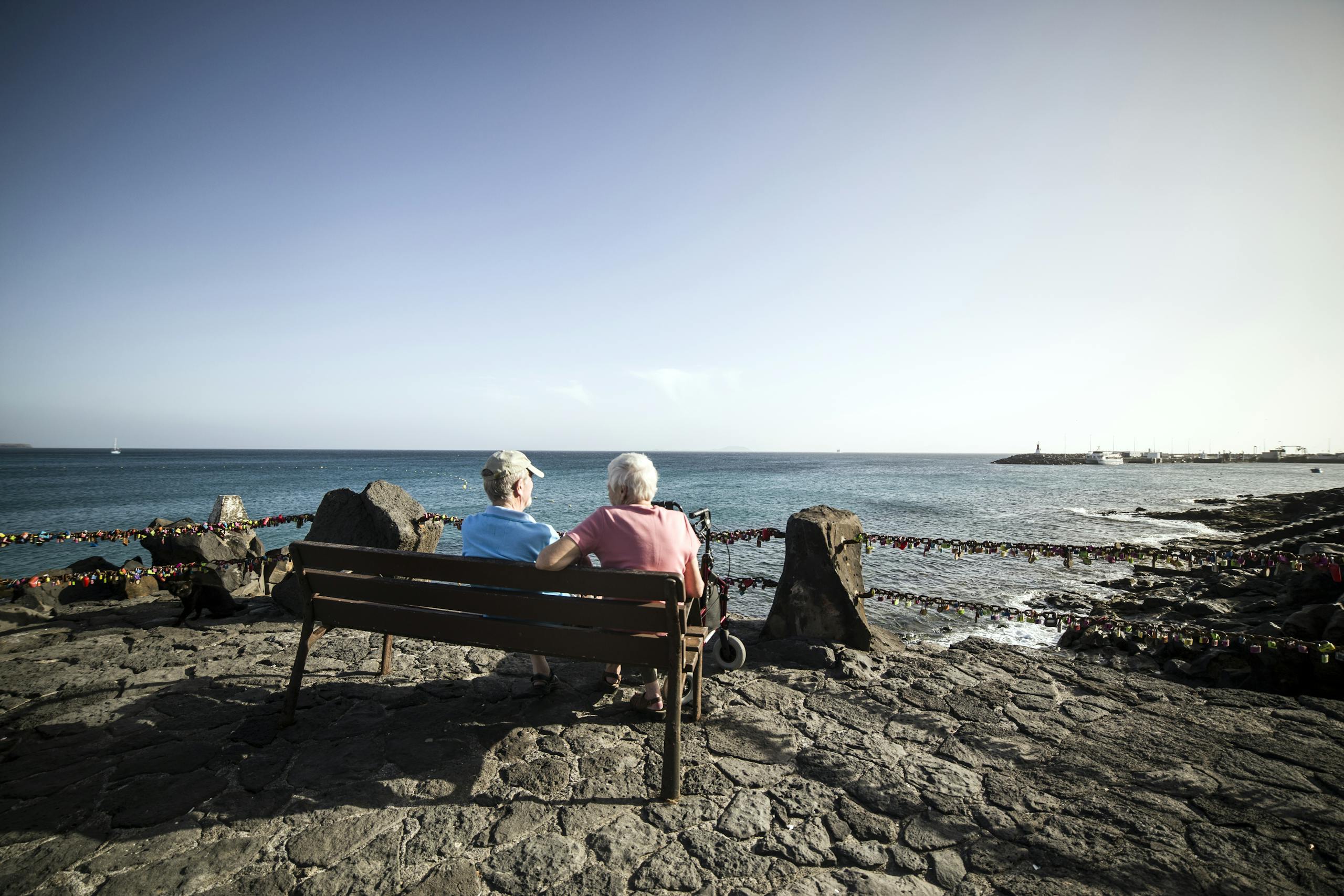 Back view full length faceless senior couple relaxing on bench on stone promenade and chatting while enjoying amazing peaceful seascape during sunny weather