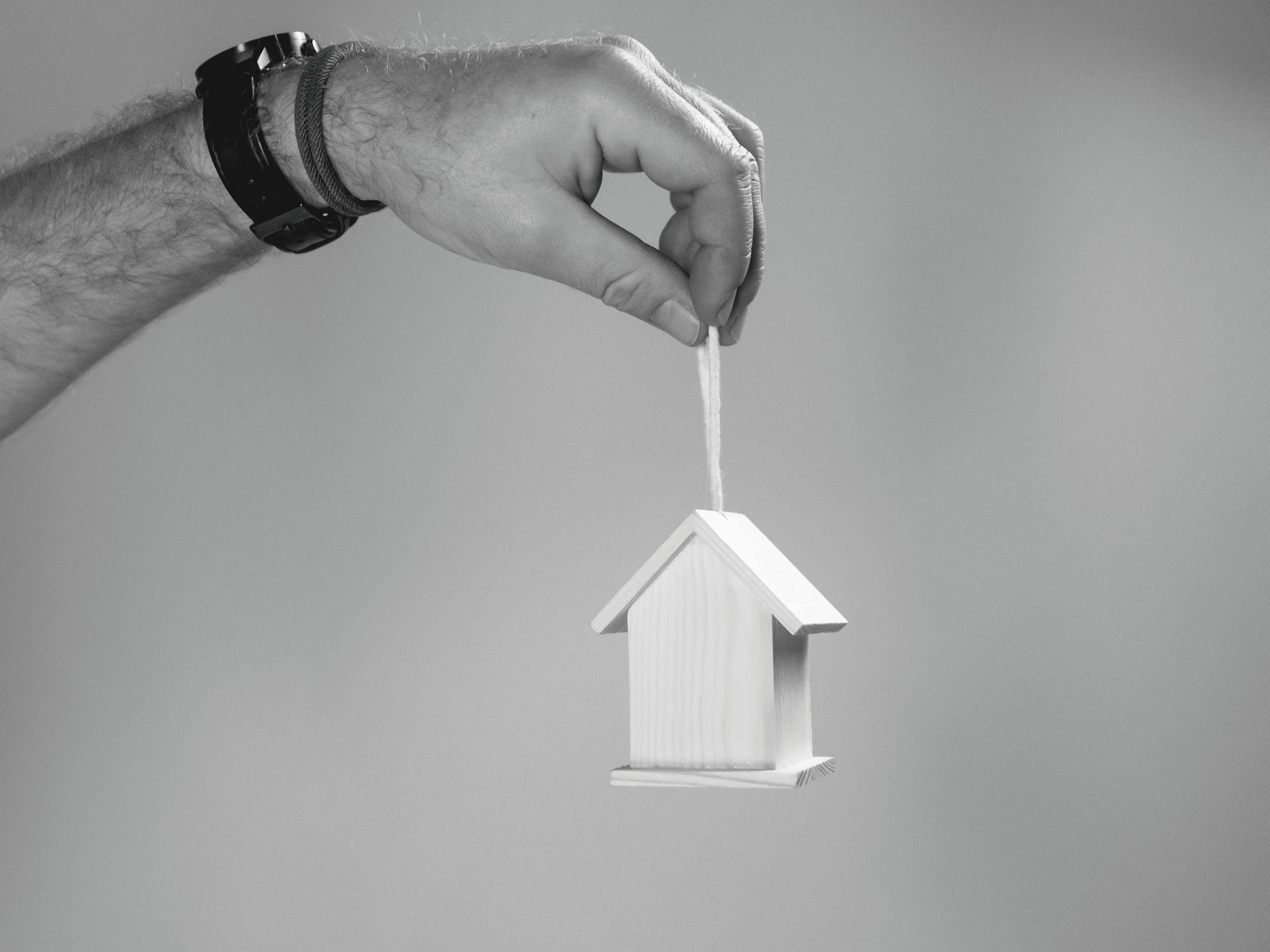 Black and white photo of a hand holding a miniature wooden house by a string, symbolizing real estate.