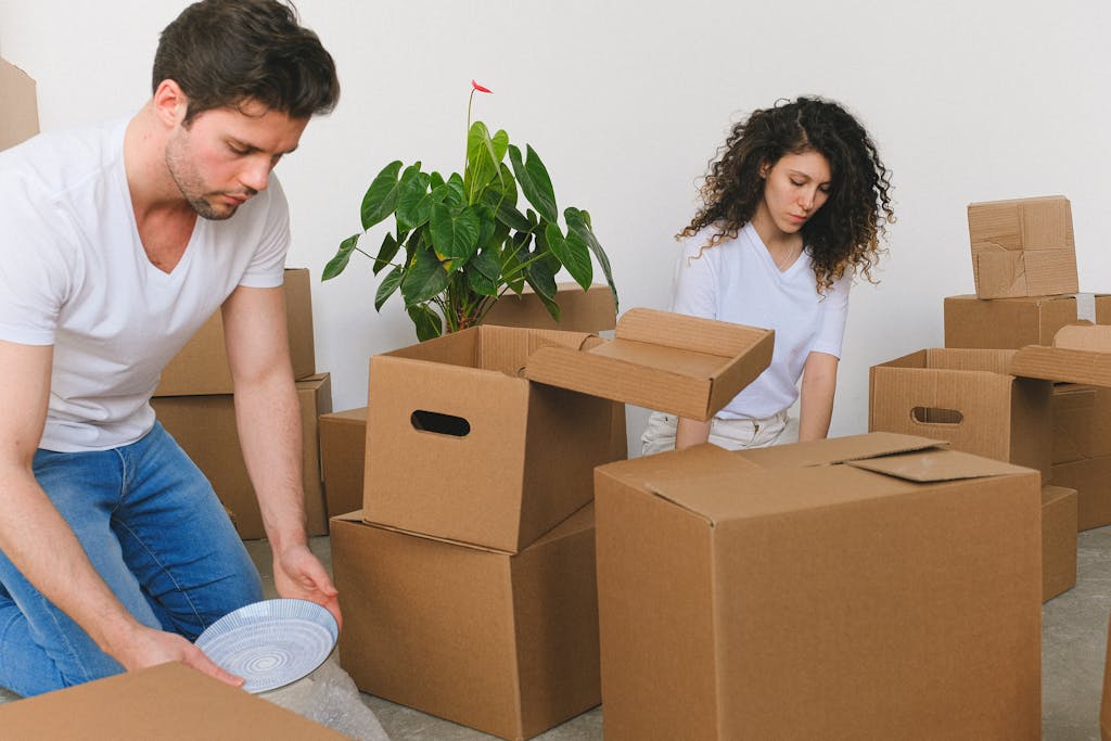 A young couple packing boxes as they move into a new apartment. Cardboard boxes and a plant are visible.